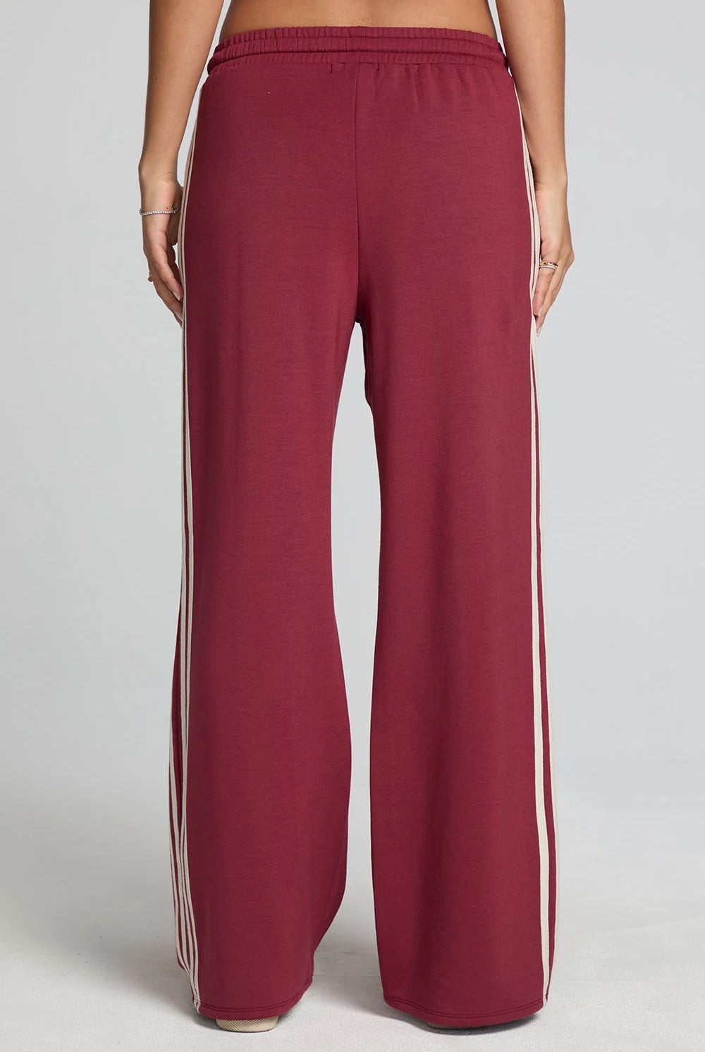 A person shown from the back wearing wide-leg sweatpants in a deep burgundy color with white stripes down the sides, against a light gray studio background.