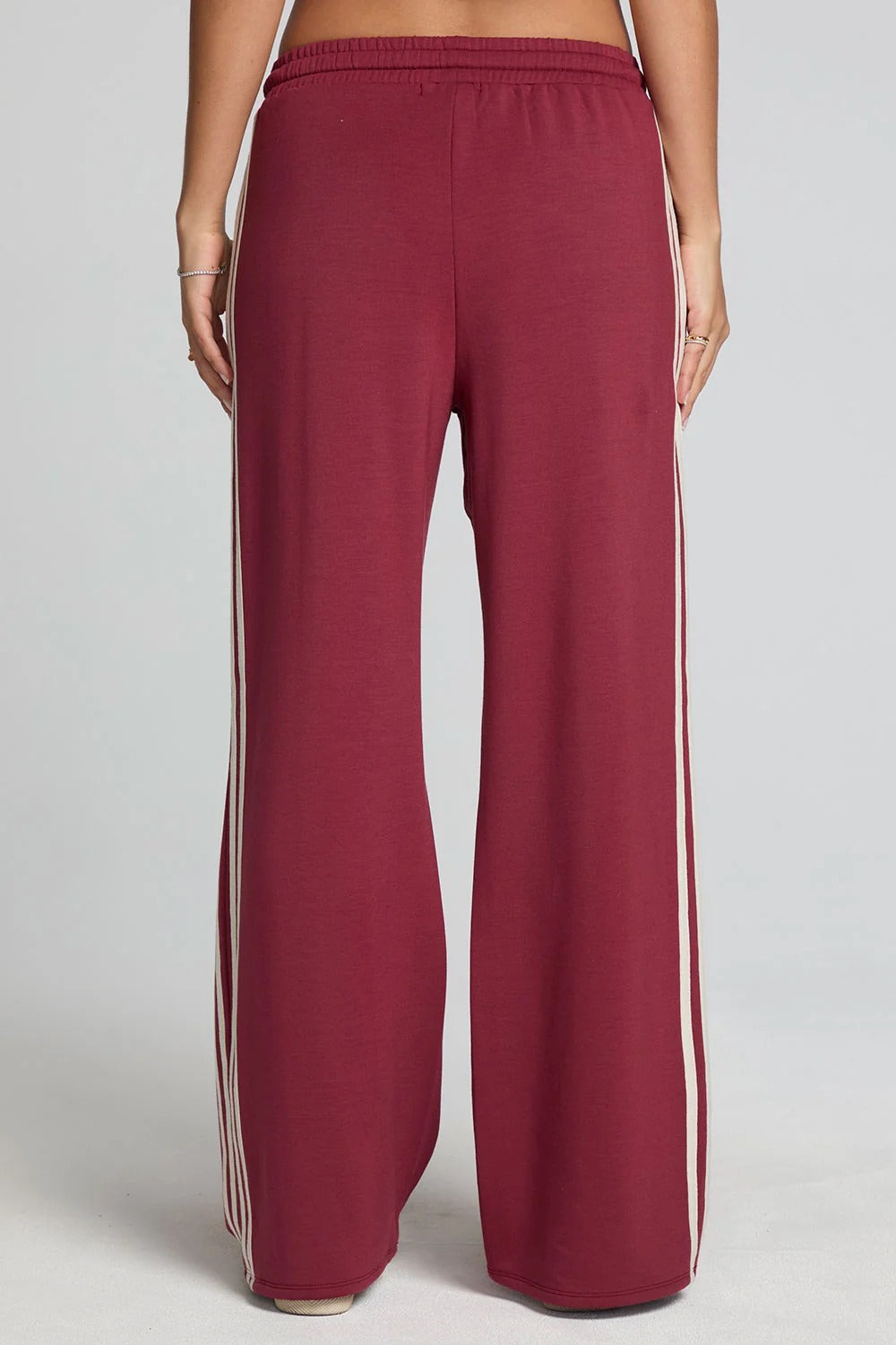 A person shown from the back wearing wide-leg sweatpants in a deep burgundy color with white stripes down the sides, against a light gray studio background.