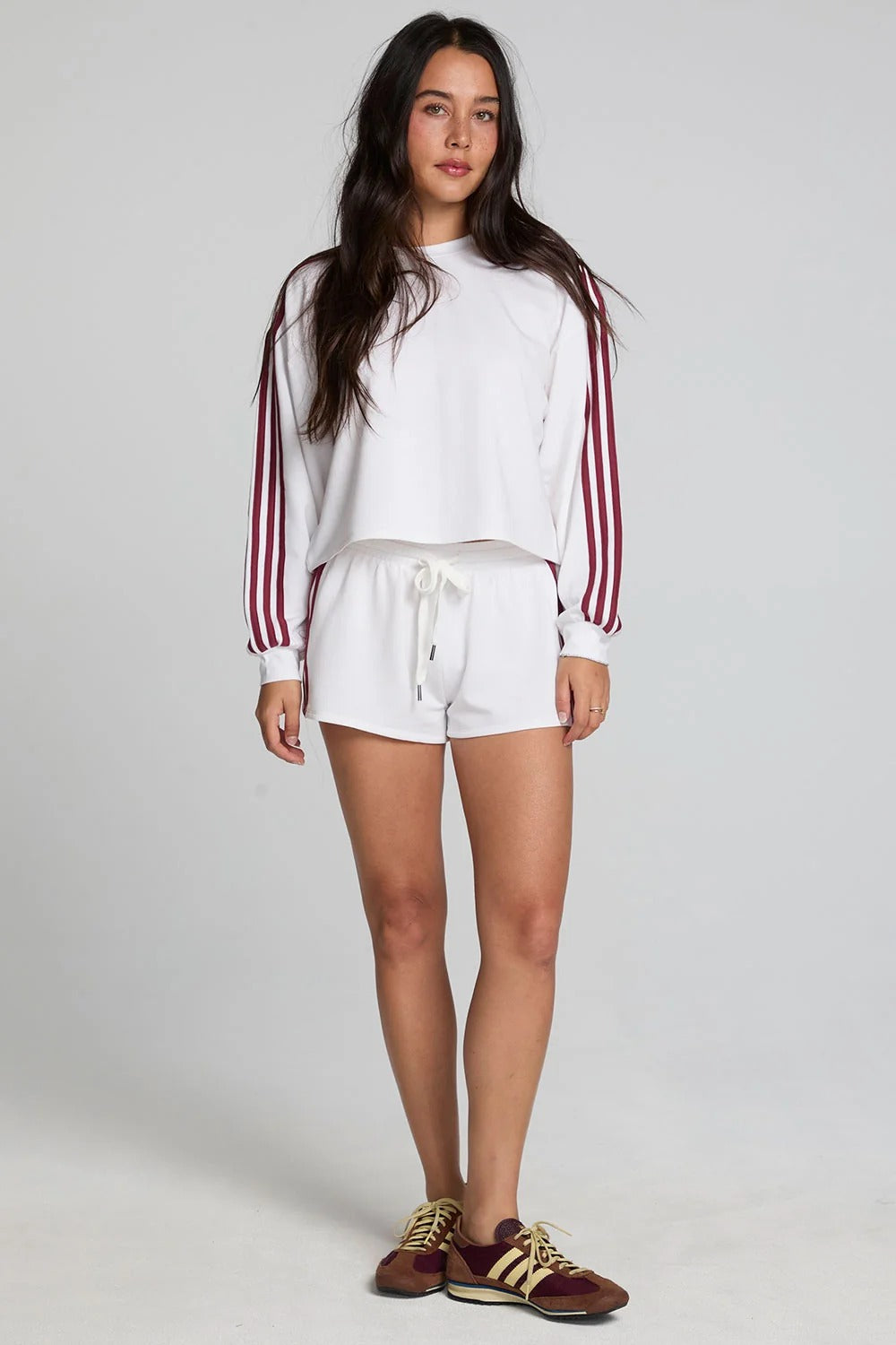 A woman modeling a white cropped sweatshirt with burgundy stripes down the sleeves and matching white shorts, paired with brown sneakers, against a light gray studio background.