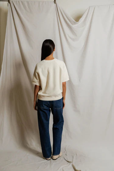 Person wearing a white shirt and blue jeans standing in front of a plain white curtain.