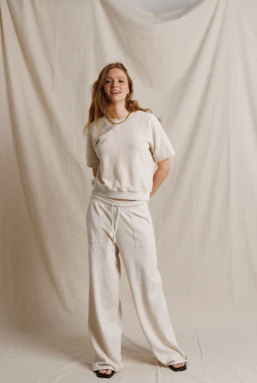 A young woman stands against a draped cream fabric backdrop, wearing a matching cream lounge set consisting of a short-sleeve cropped sweatshirt and wide-leg sweatpants.