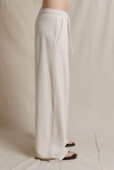 A person wearing loose, wide-leg white drawstring pants with a side pocket, shown from the waist down against a neutral beige fabric backdrop.