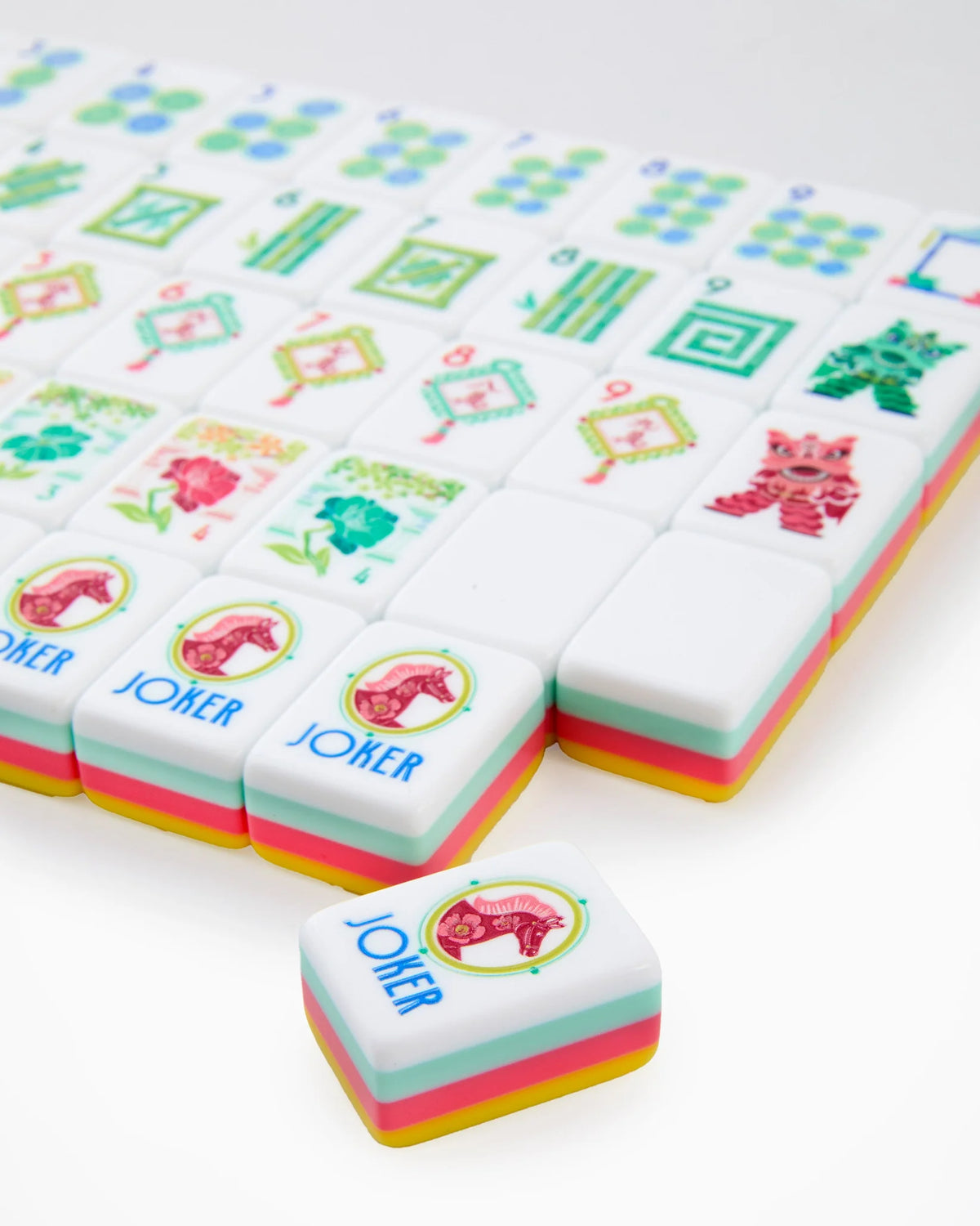 A close-up of a set of Mahjong-style tiles on a white background, with one tile in front showing the Joker symbol and pastel pink, yellow, and mint green layered edges.