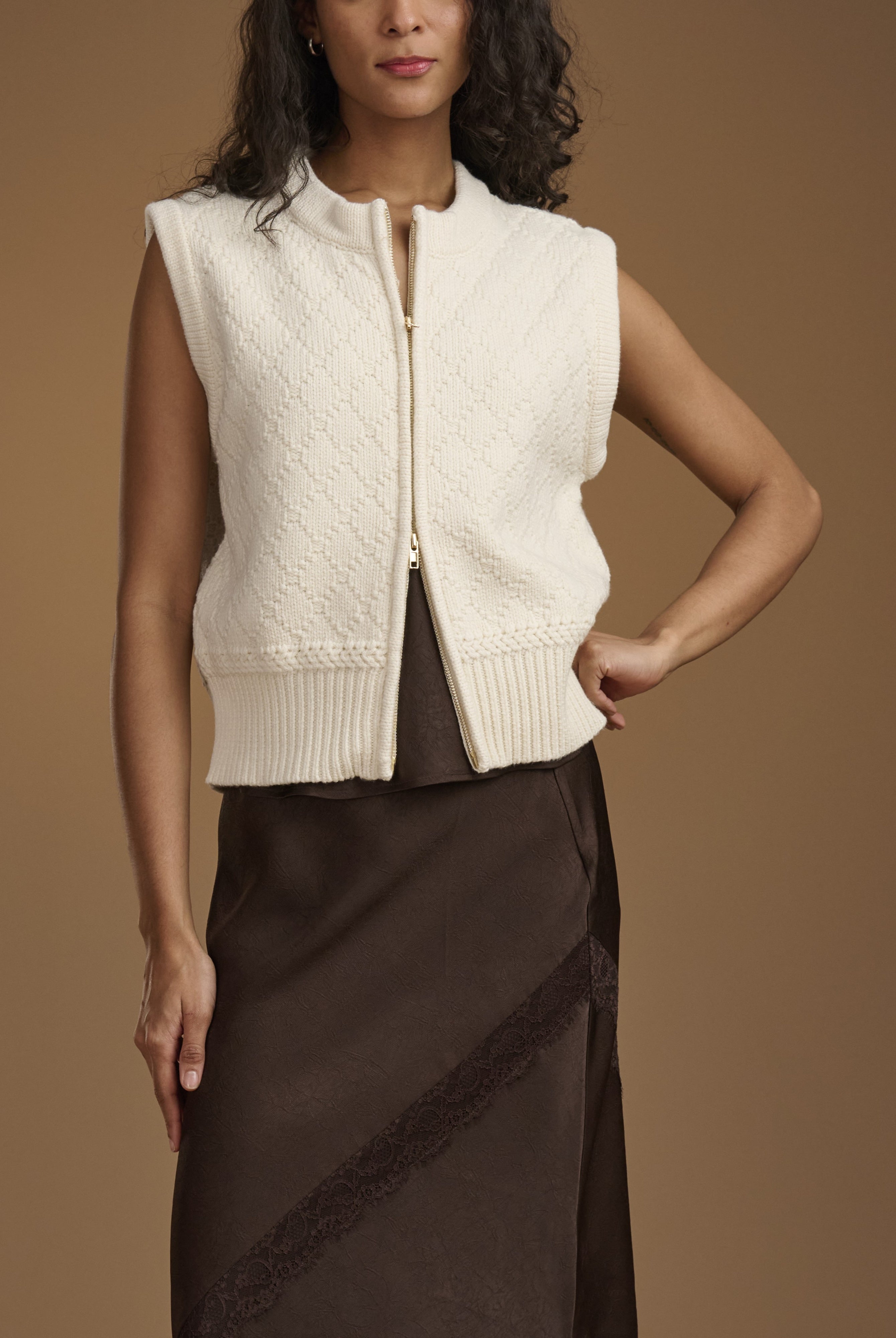 A woman models a sleeveless cream cable-knit zip-front vest over a dark brown skirt, set against a solid warm brown studio background.
