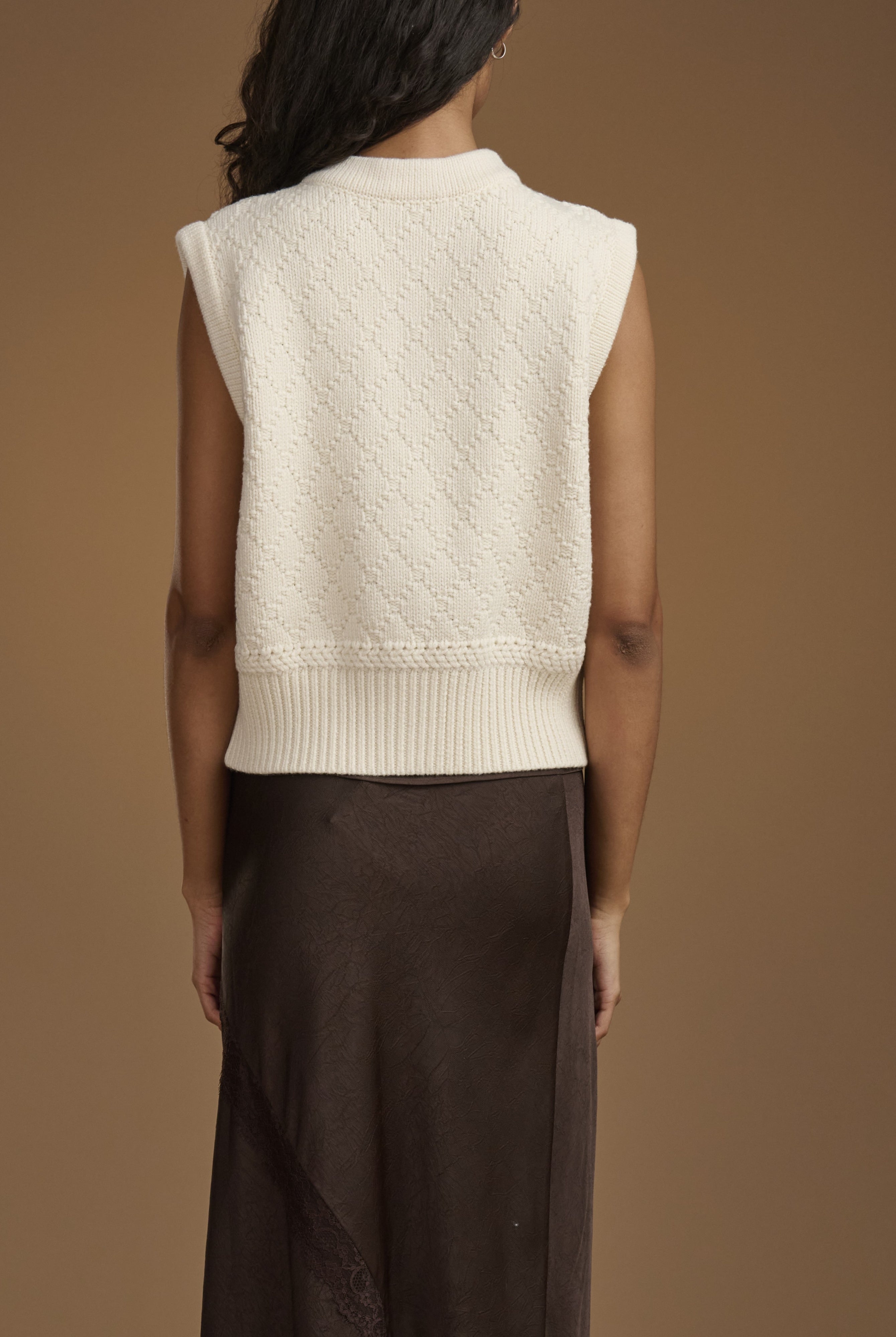 Back view of a model wearing a cream-colored sleeveless knit sweater vest over a dark brown skirt, set against a solid warm brown studio background.