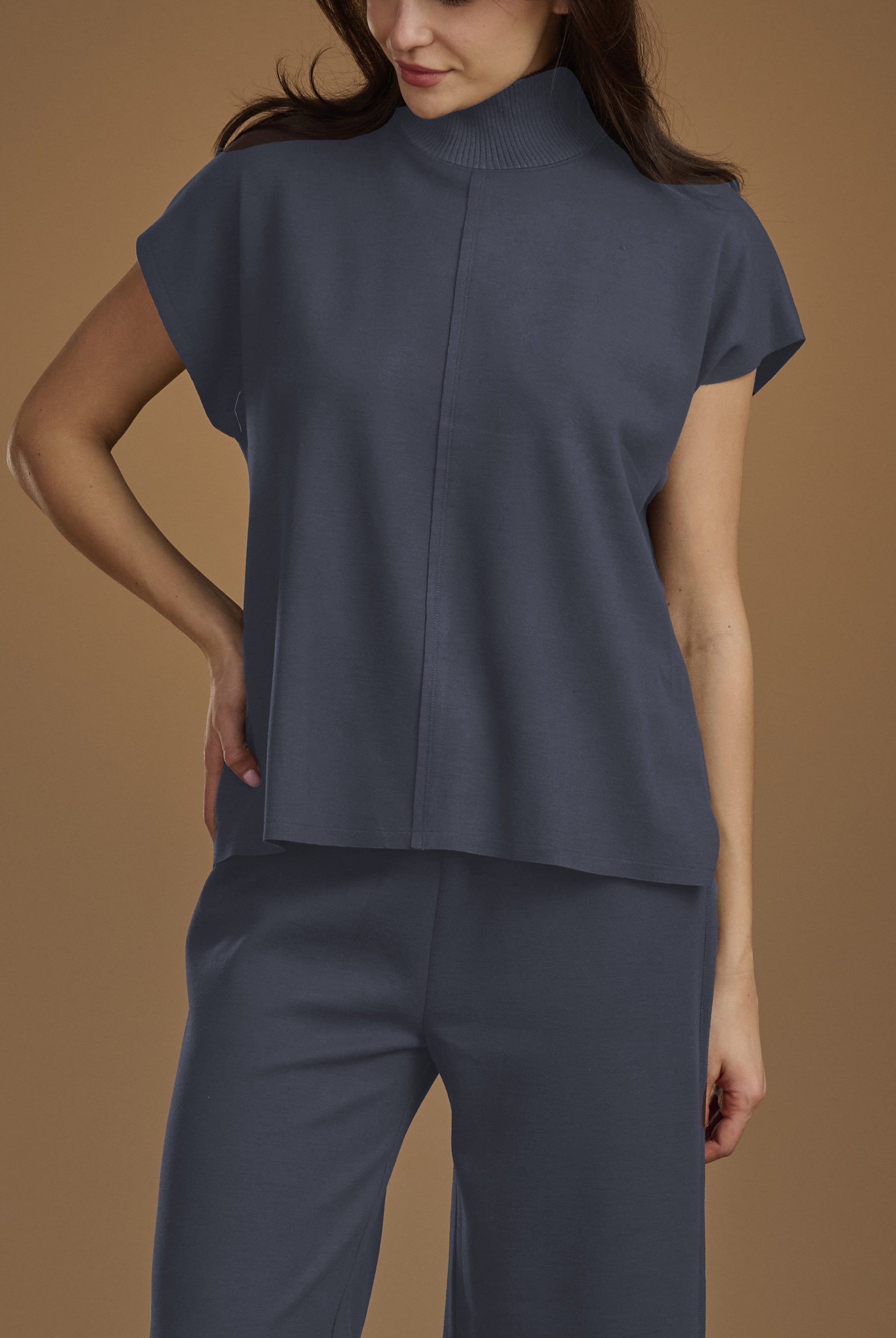 A woman wearing a slate blue mock-neck short-sleeve top and matching wide-leg trousers, posed against a solid warm brown studio background.