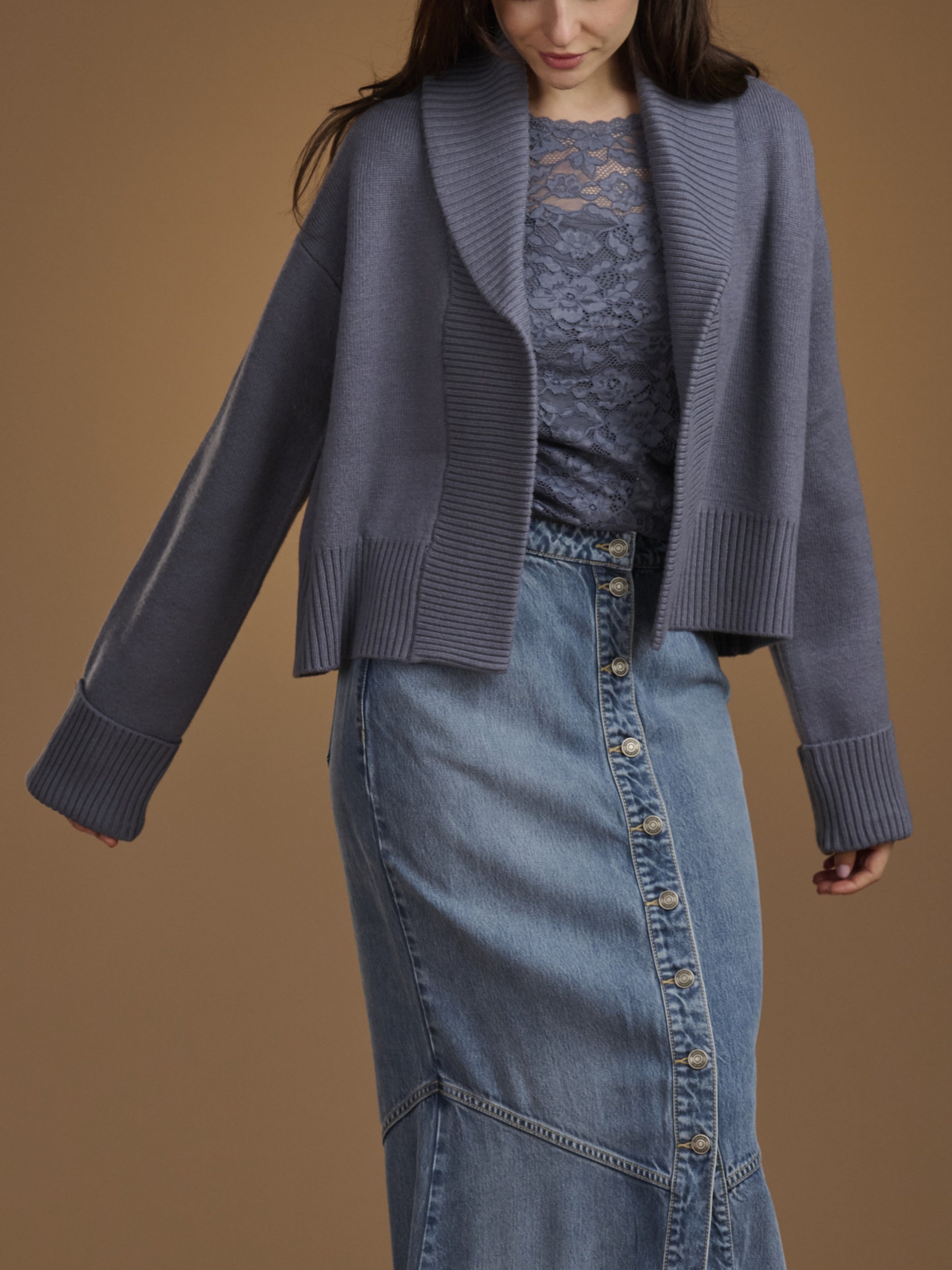 A woman wearing a slate blue open-front knit cardigan over a matching lace top and a blue denim skirt, photographed against a solid warm brown studio background.