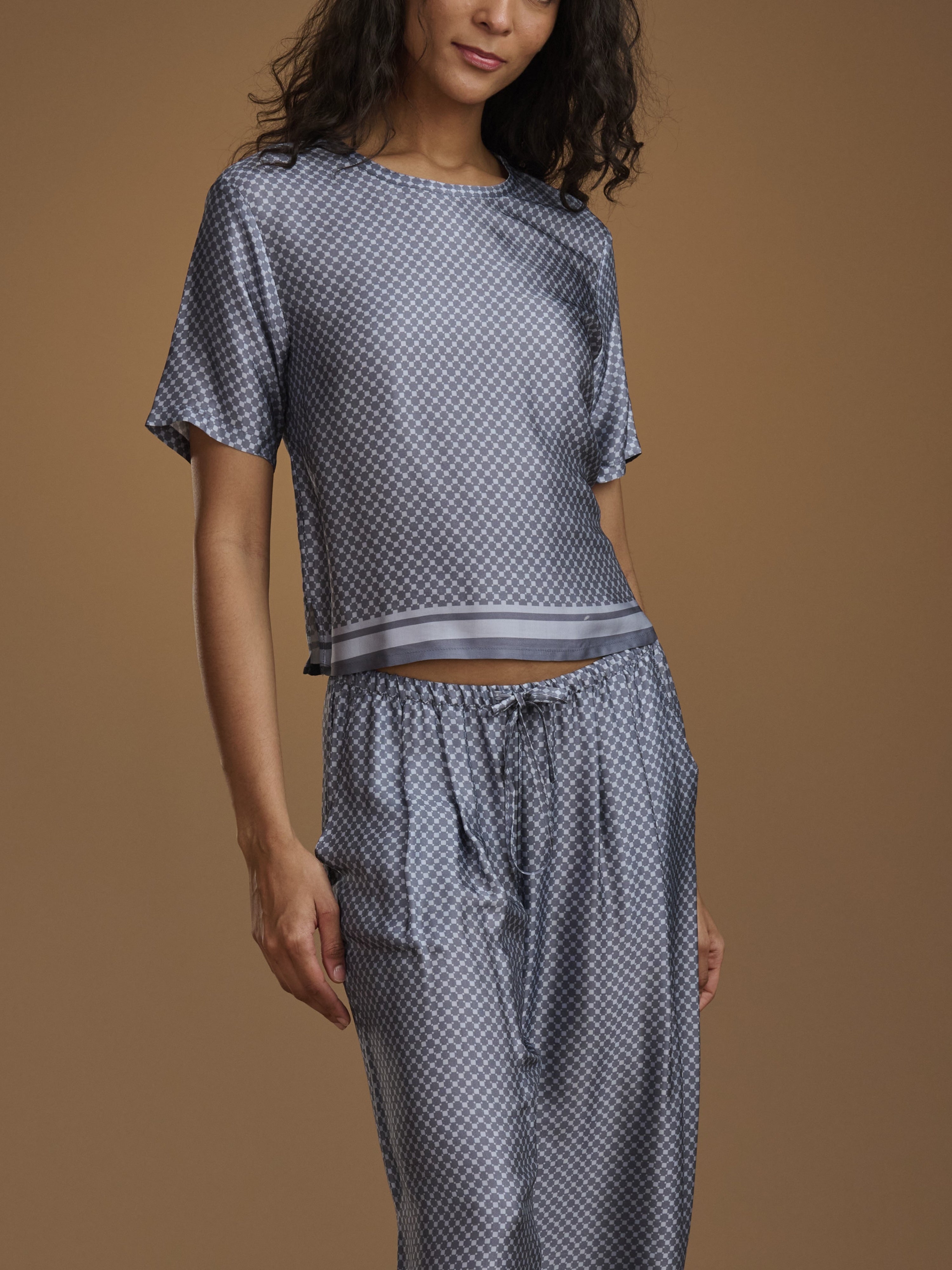 A woman wearing a matching two-piece loungewear set with a blue and white geometric pattern, consisting of a short-sleeve crop top and drawstring pants, against a solid brown studio background.