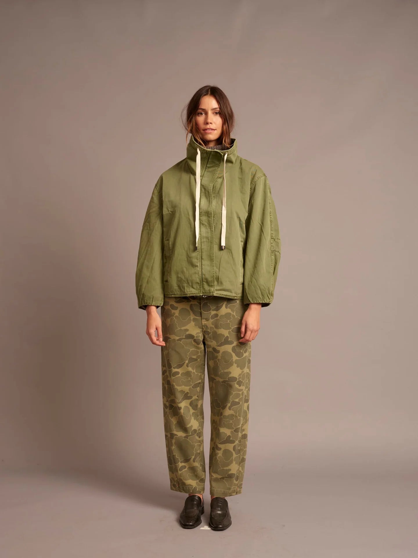 A model stands against a plain taupe studio background wearing an olive green hooded jacket with cream drawstrings and olive green camouflage pants.