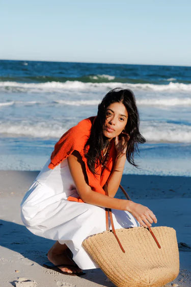 Woman in orange top and white pants holding a straw bag on a beach with ocean waves in the background
