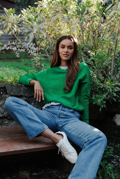 Woman in a green sweater and blue jeans sitting outdoors among plants