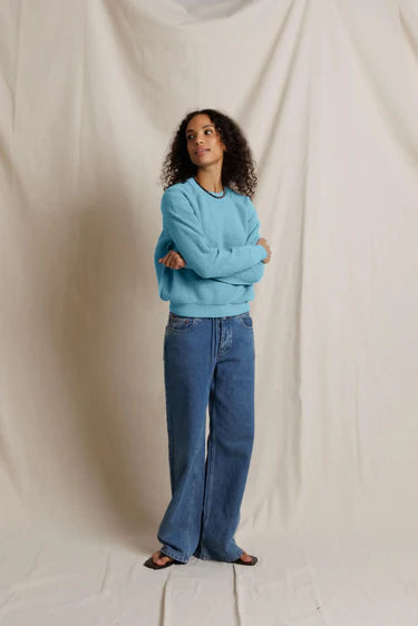 A woman stands against a draped off-white fabric backdrop, wearing a light blue crewneck sweatshirt and wide-leg blue jeans.