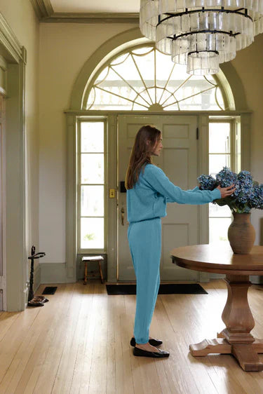 A woman in a light blue matching two-piece outfit stands in a bright, elegant entryway, reaching toward a vase of blue hydrangeas on a round wooden table. The scene features a large arched window, a crystal chandelier, and light hardwood floors.
