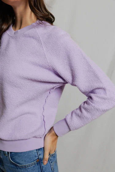 Person wearing a purple textured sweater and blue jeans on a plain background