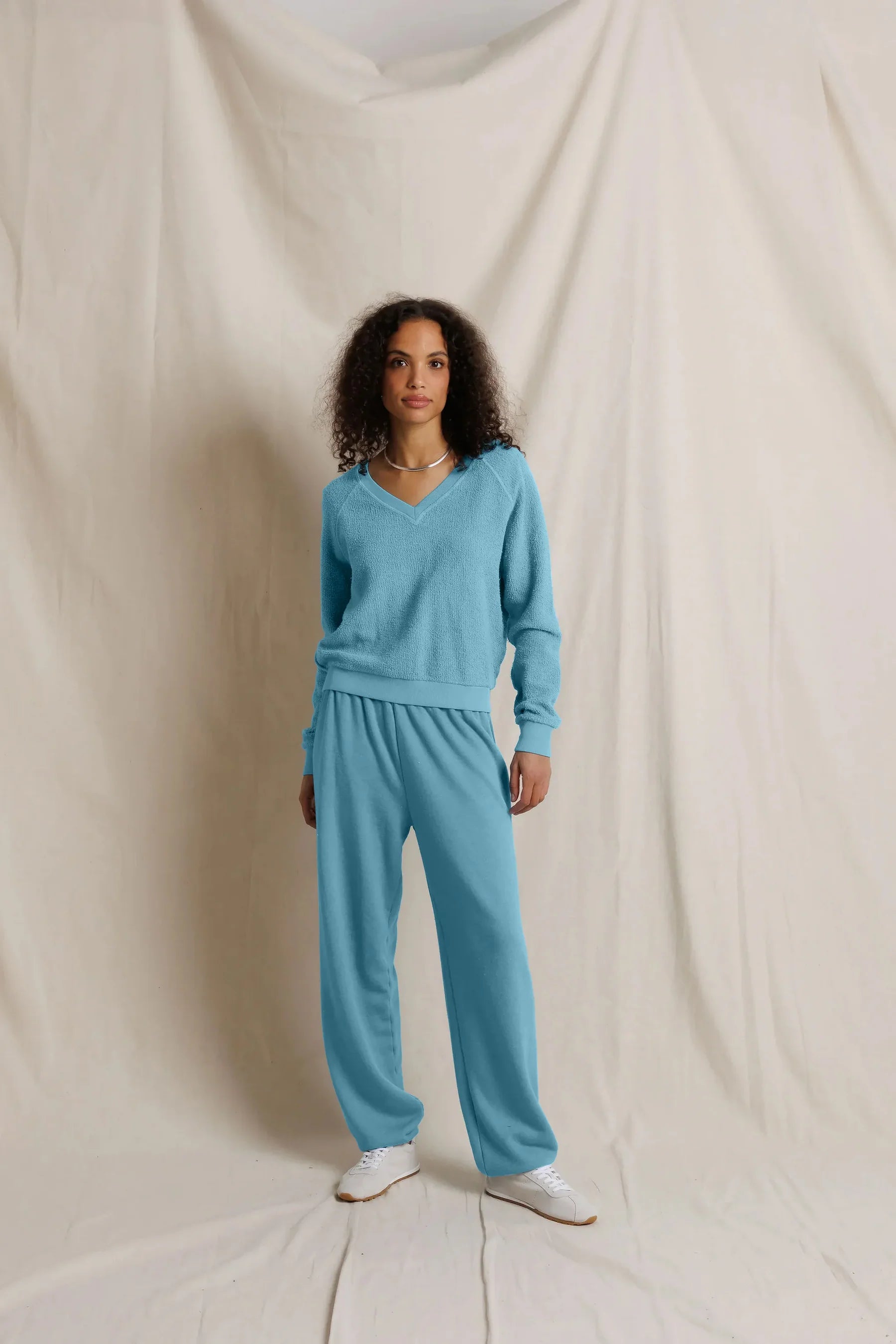 A model stands against a draped cream fabric backdrop wearing a matching light blue knit sweater and sweatpants set, with white sneakers.