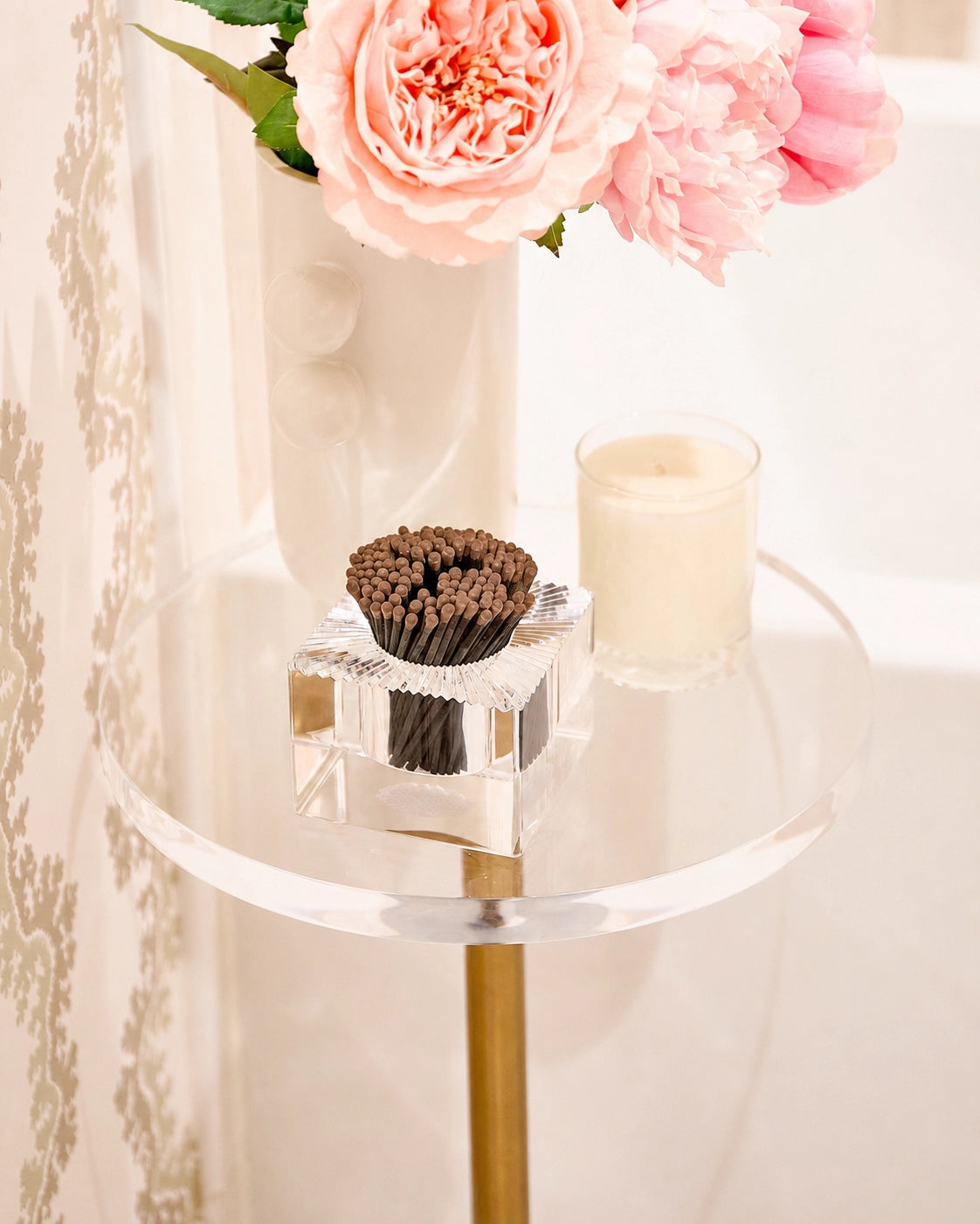 A clear acrylic side table with a gold metal stand holding a small white candle and a square glass holder filled with dark brown matches. A white vase with pink peonies sits behind the table against a softly lit interior background.