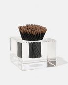 A set of black matchsticks with brown tips arranged inside a clear acrylic square container on a white background.