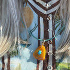 A close-up of a person wearing a turquoise cord necklace with a bright yellow-orange teardrop-shaped pendant featuring a small turquoise stone in the center. The person is wearing a light blue and brown patterned top with white buttons.