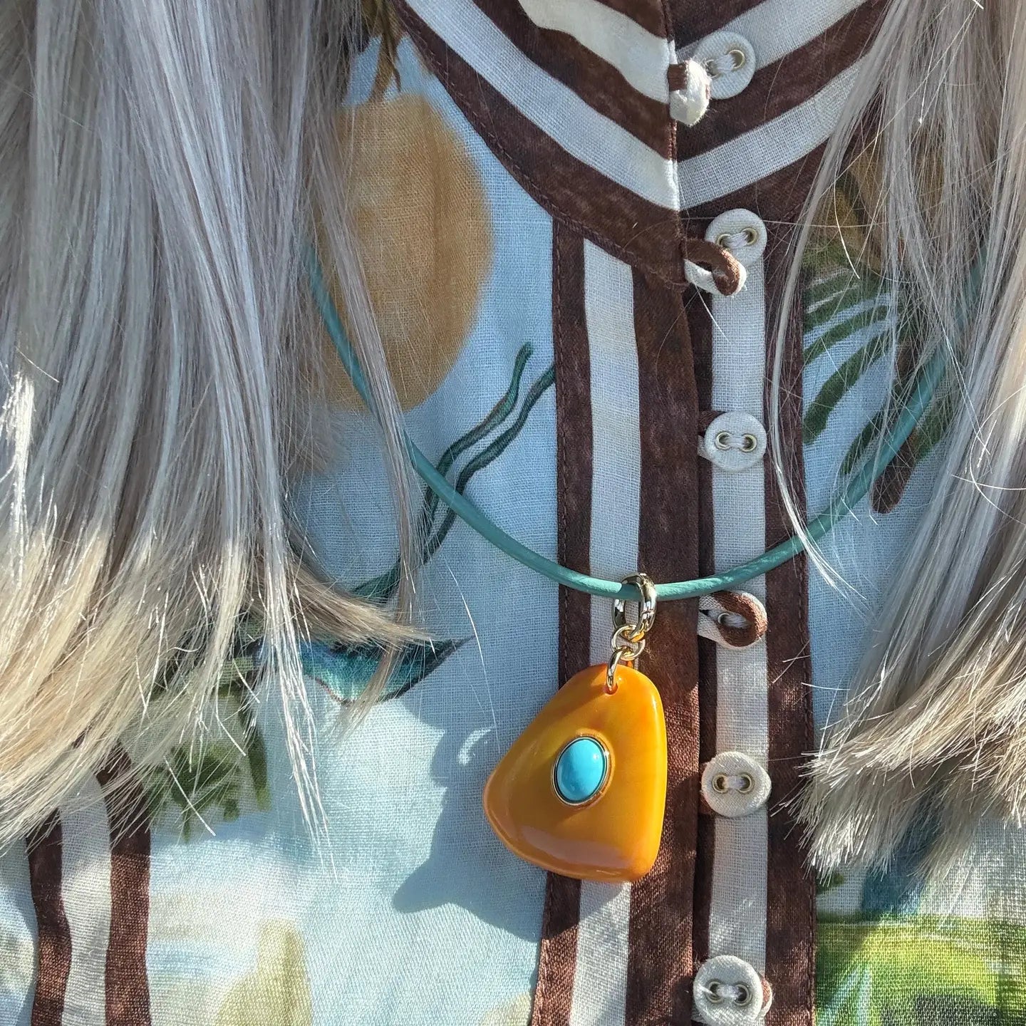 A close-up of a person wearing a turquoise cord necklace with a bright yellow-orange teardrop-shaped pendant featuring a small turquoise stone in the center. The person is wearing a light blue and brown patterned top with white buttons.