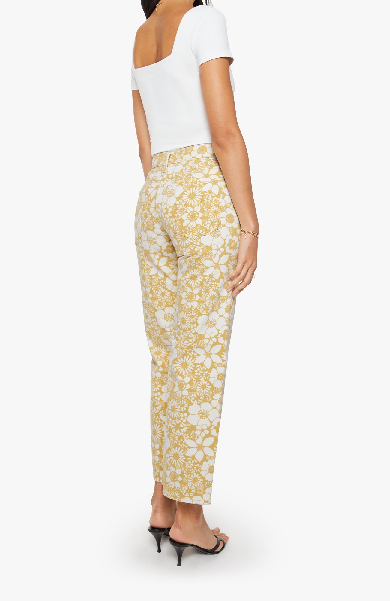 A model shown from the back wearing yellow and white floral print trousers, a white off-the-shoulder top, and black high-heeled sandals against a light gray background.