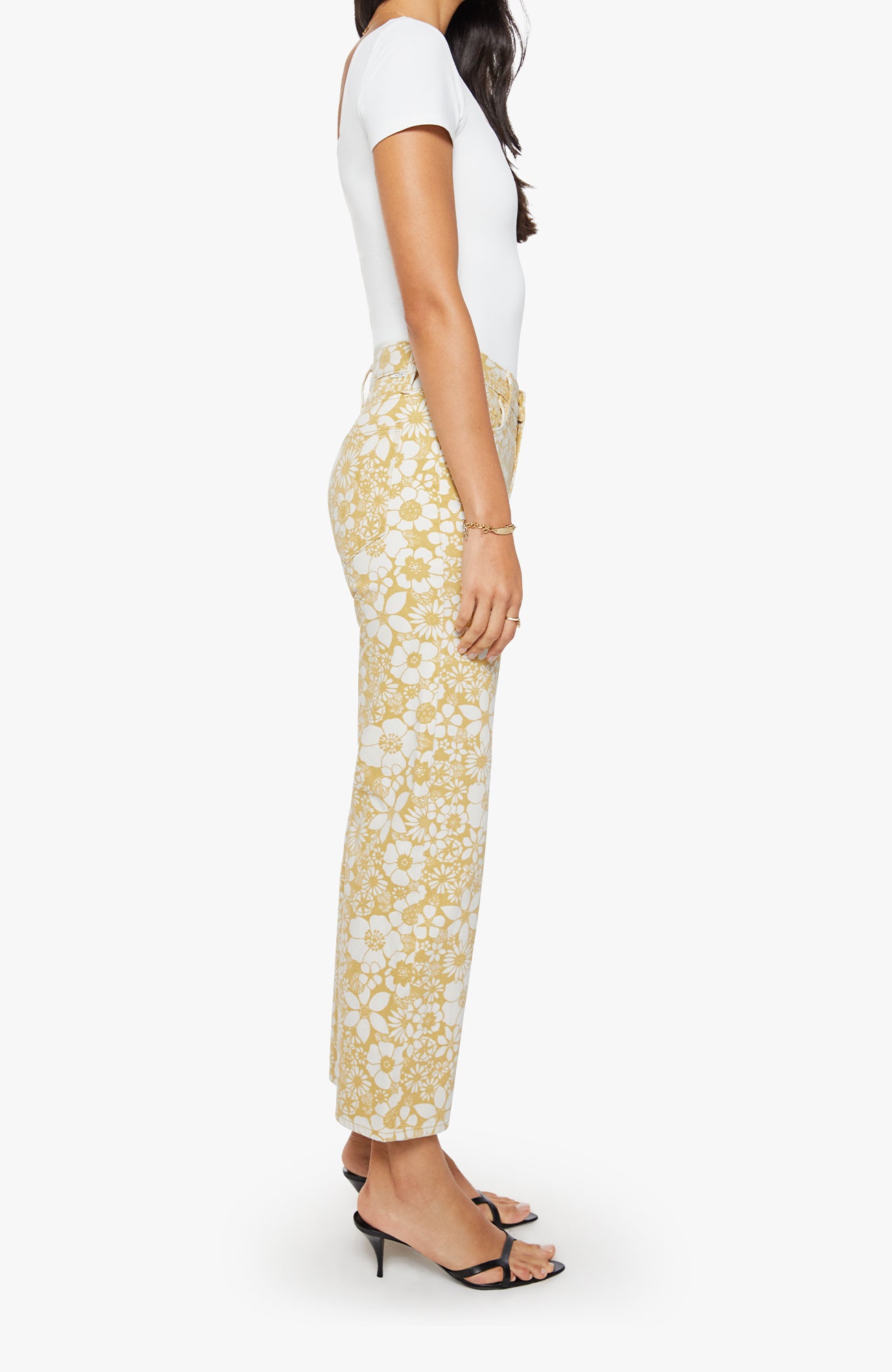 A side-profile view of a model wearing high-waisted, straight-leg trousers with a white and yellow floral lace pattern, paired with a white top and black heeled sandals, against a light gray background.