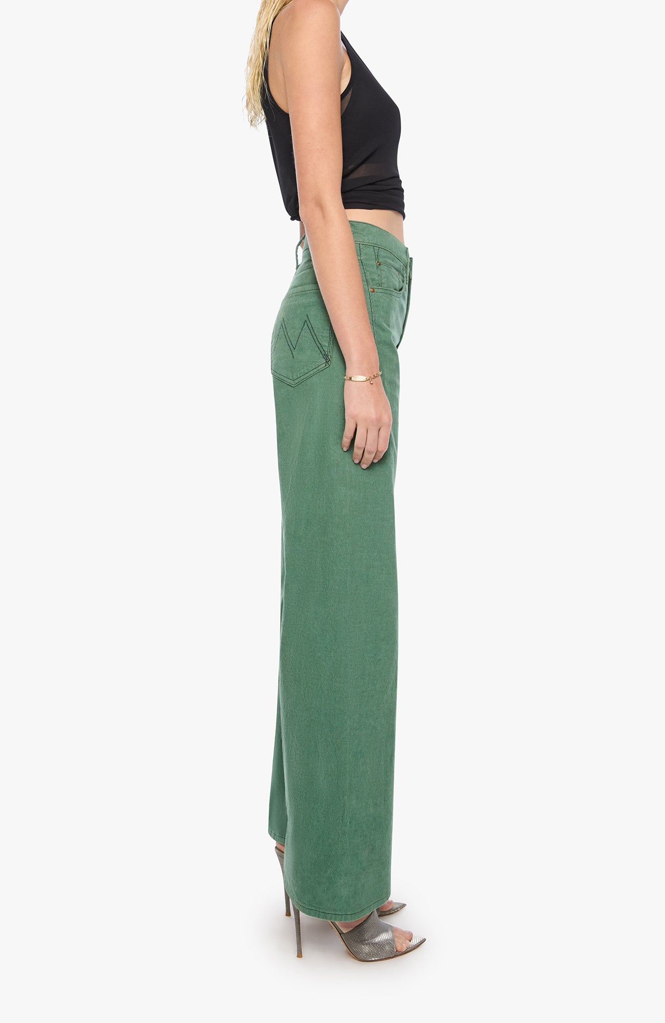 A side-profile view of a model wearing high-waisted, wide-leg sage green denim pants with a black sleeveless crop top and metallic high-heeled sandals, set against a plain white background.