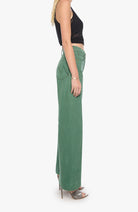 A side-profile view of a model wearing high-waisted, wide-leg sage green denim pants with a black sleeveless crop top and metallic high-heeled sandals, set against a plain white background.