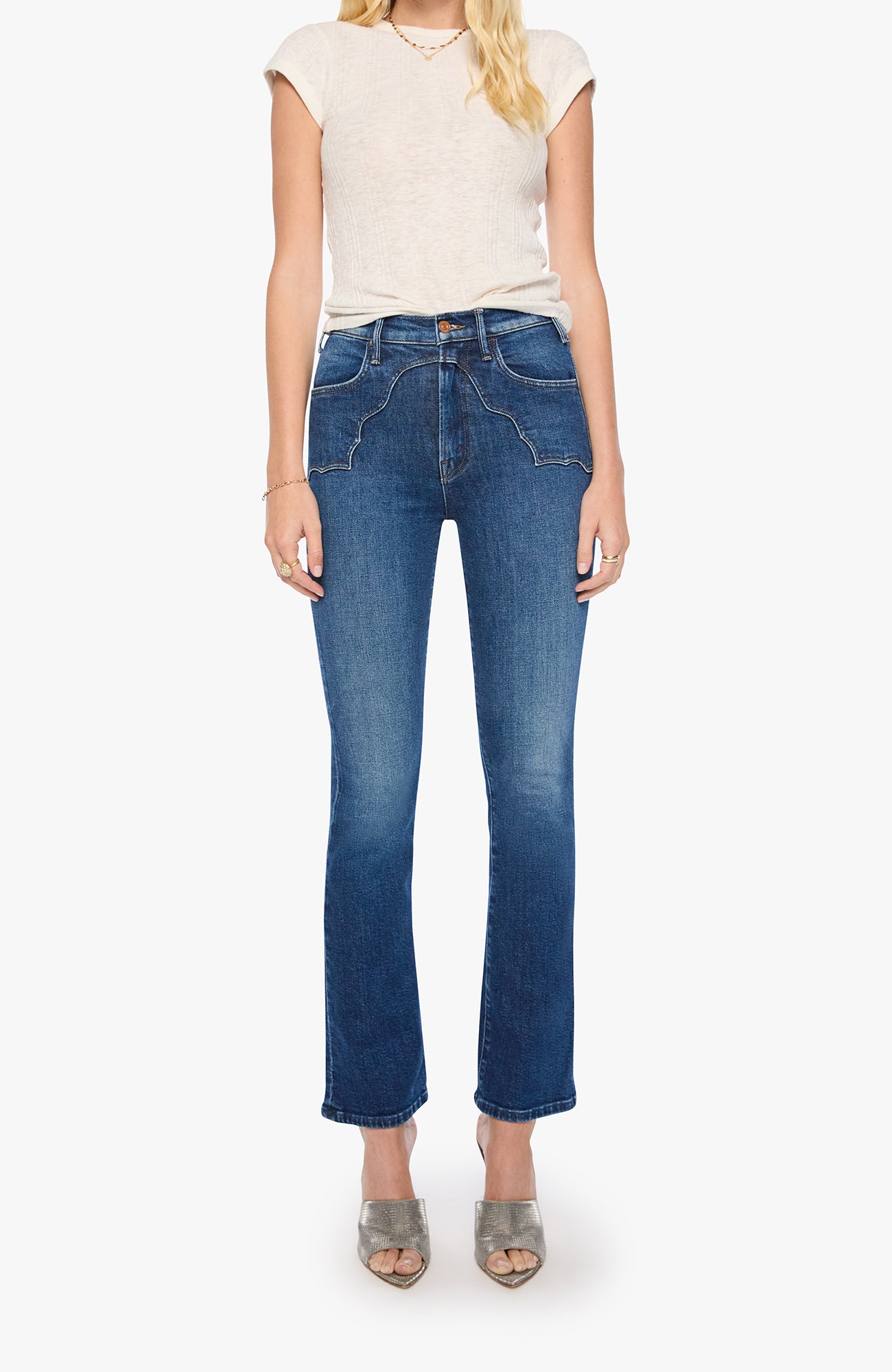 A model wearing a pair of high-waisted, straight-leg blue jeans with a faded wash, styled with a cream-colored sleeveless top and gray heeled sandals against a plain white background.