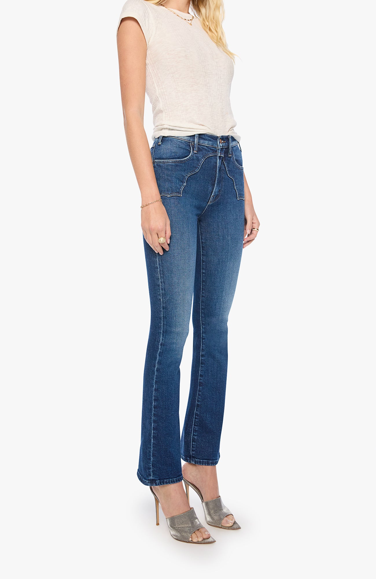 A model wearing a pair of medium-wash blue flare jeans, paired with a cream sleeveless top and gray high-heeled sandals, shown against a plain white background.