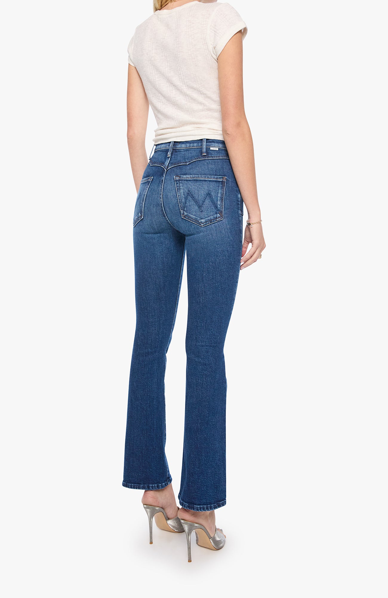 A woman shown from the back wearing blue flare jeans, a cream short-sleeve top, and metallic high heels against a plain light background.