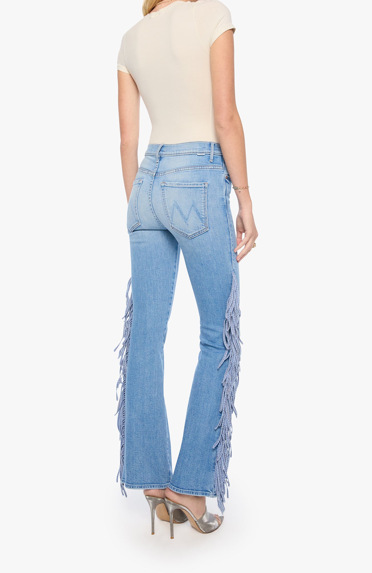 A model shown from the back wearing light-wash blue denim flare jeans with long fringe detailing down the outer sides, paired with a cream top and metallic heels.
