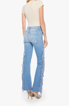 A model shown from the back wearing light-wash blue denim flare jeans with long fringe detailing down the outer sides, paired with a cream top and metallic heels.