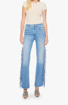 A model wearing light blue high-waisted flare jeans with long fringe detailing down the outer legs, paired with a cream short-sleeve top and metallic silver heels, against a plain light gray background.