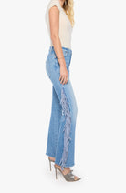 A side-profile view of a model wearing light blue wide-leg denim jeans with long fringe detailing running down the outer side of each leg, paired with a cream sleeveless top and metallic gray high-heeled sandals against a plain white background.