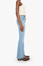 A side-profile view of a model wearing light-wash blue flared jeans, a cream ribbed tank top, and brown leather boots against a plain white studio background.