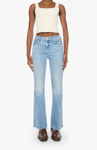 A model wearing light-wash blue high-rise flare jeans, paired with a cream-colored crop top and brown leather shoes, standing against a plain white studio background.