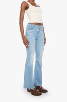 A model wearing light-wash blue flare jeans, a cream ribbed tank top, and brown leather shoes, standing against a plain white studio background.