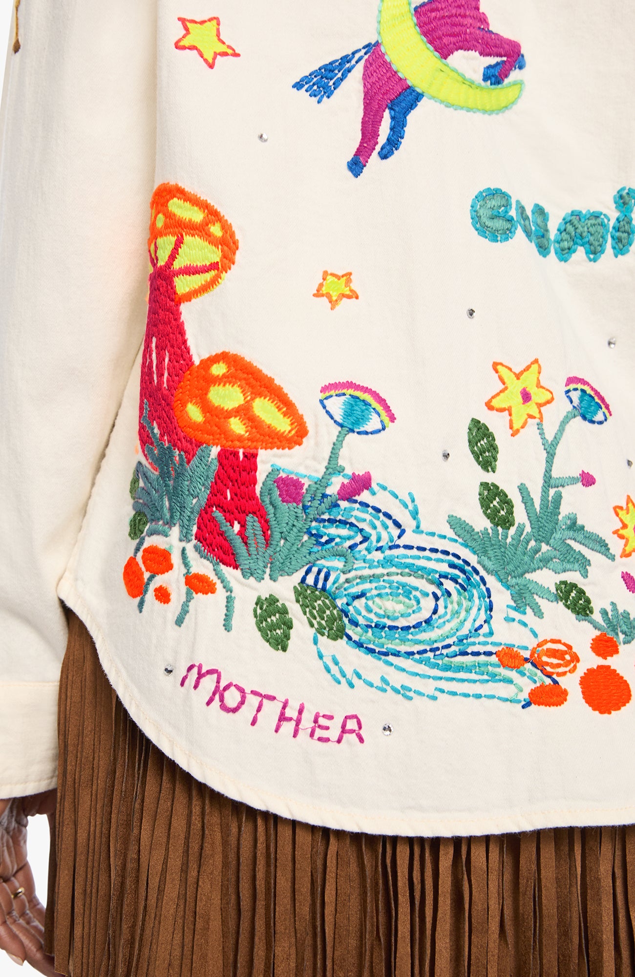 Close-up of a cream-colored embroidered garment featuring colorful whimsical embroidery of mushrooms, flowers, a stream, stars, and the word 'MOTHER'.