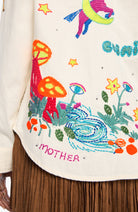 Close-up of a cream-colored embroidered garment featuring colorful whimsical embroidery of mushrooms, flowers, a stream, stars, and the word 'MOTHER'.