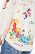 Close-up of a cream-colored embroidered shirt worn with blue jeans. The shirt features colorful embroidery of a pink unicorn on a crescent moon, orange and red mushrooms, stars, and the word 'MOTHER' in pink.