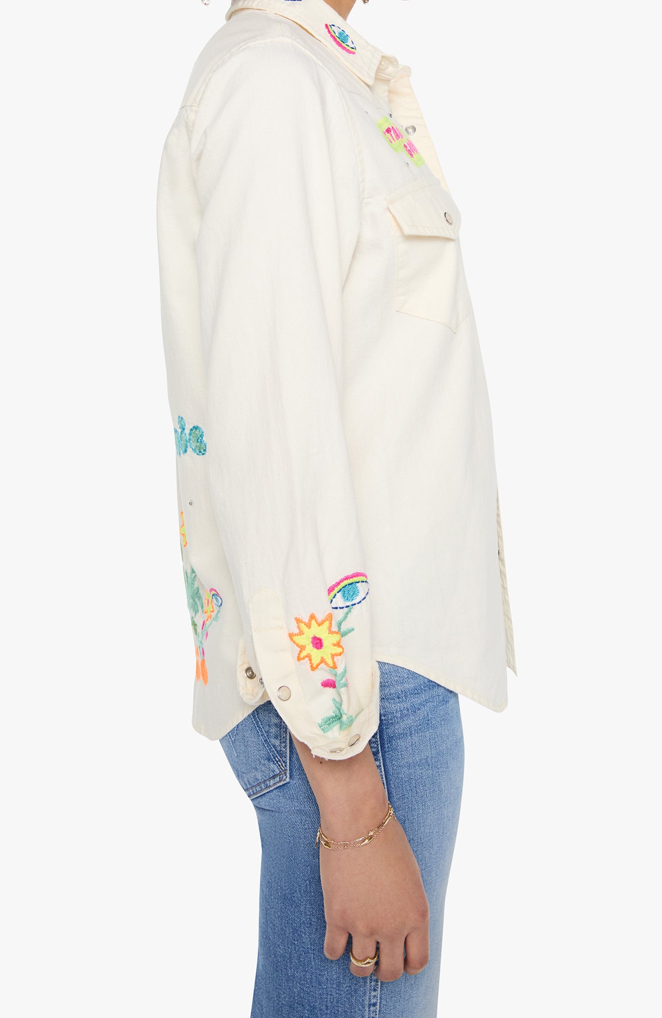 Side view of a person wearing an off-white button-up shirt with colorful embroidered floral and insect motifs, paired with blue jeans against a plain white background.