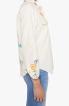 Side view of a person wearing an off-white button-up shirt with colorful embroidered floral and insect motifs, paired with blue jeans against a plain white background.