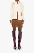A model is shown from the waist down wearing a cream-colored embroidered button-up shirt, a brown fringe mini skirt, sheer brown tights, and dark burgundy pointed-toe knee-high boots, set against a plain light gray background.