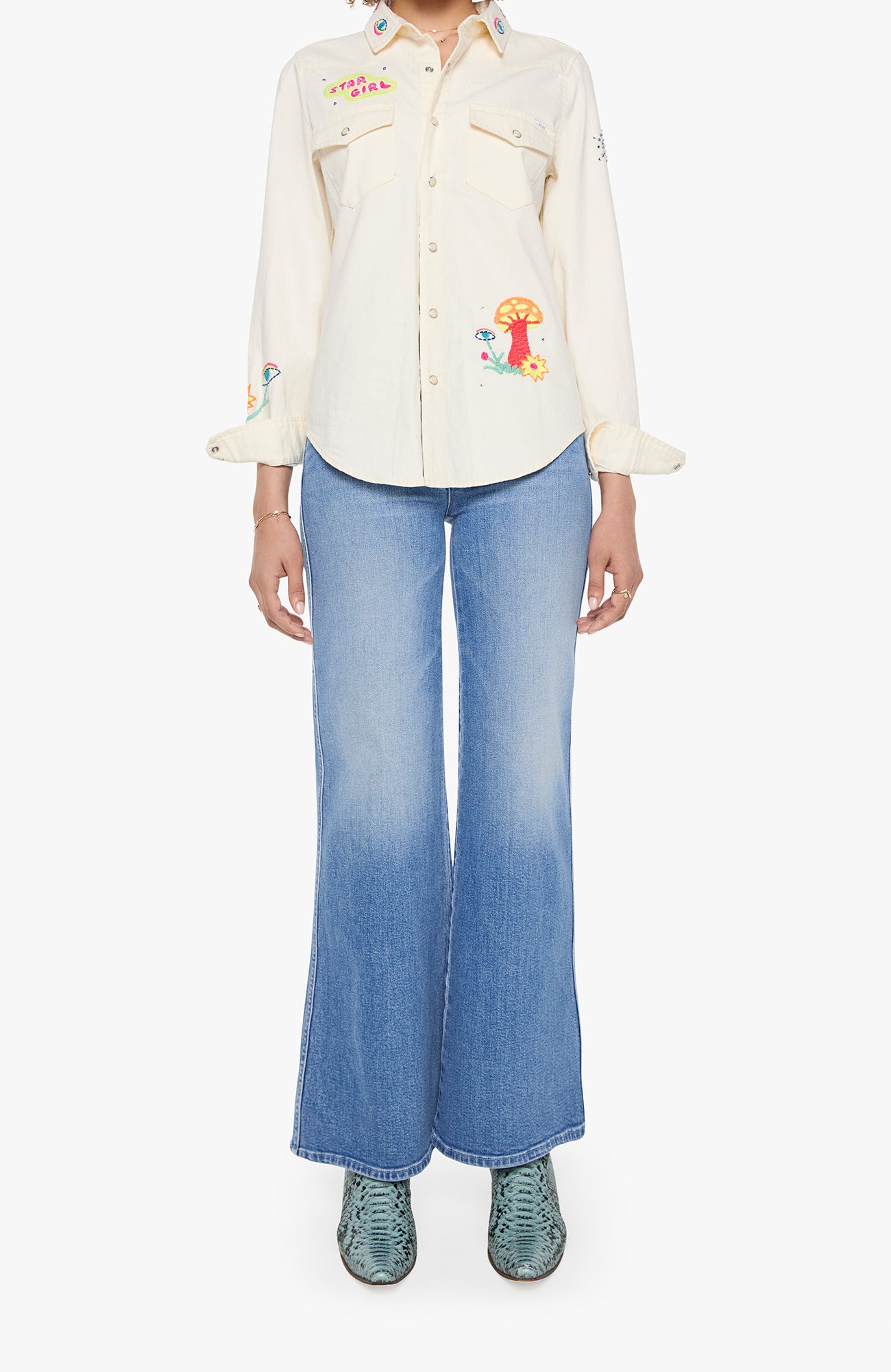 A model wearing a cream-colored embroidered button-up shirt with colorful mushroom and floral motifs, paired with wide-leg blue jeans and green snakeskin-patterned shoes, set against a plain white background.