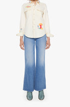 A model wearing a cream-colored embroidered button-up shirt with colorful mushroom and floral motifs, paired with wide-leg blue jeans and green snakeskin-patterned shoes, set against a plain white background.