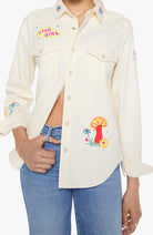 A cream-colored long-sleeve button-up shirt with colorful embroidered details, including a 'Star Girl' patch on the chest and a mushroom design on the lower front, worn by a model with blue jeans.