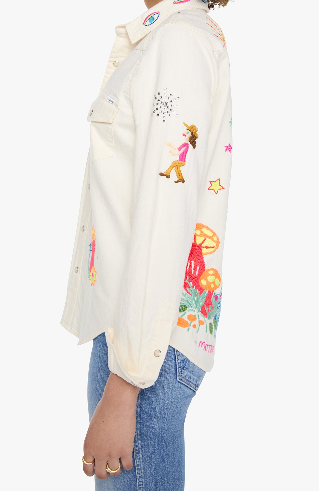 A side-profile view of a person wearing a cream-colored denim jacket with colorful cartoon and floral embroidery, paired with blue jeans, against a plain light gray background.