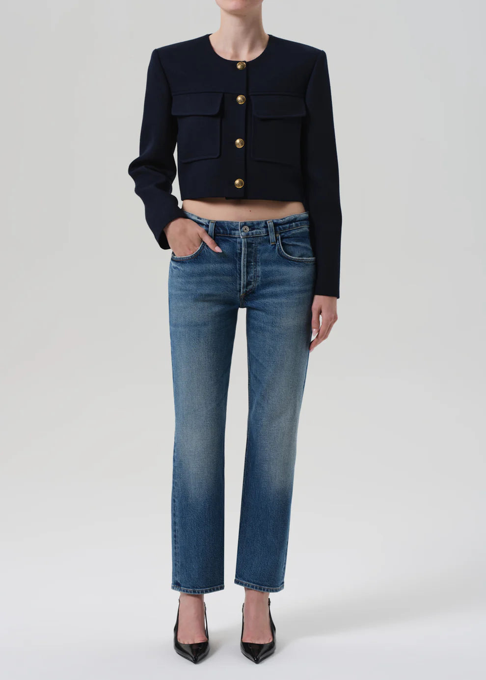 Person wearing a black cropped jacket with gold buttons and blue jeans on a white background