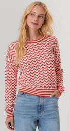 Woman wearing a red and white zigzag patterned sweater and blue jeans on a plain background