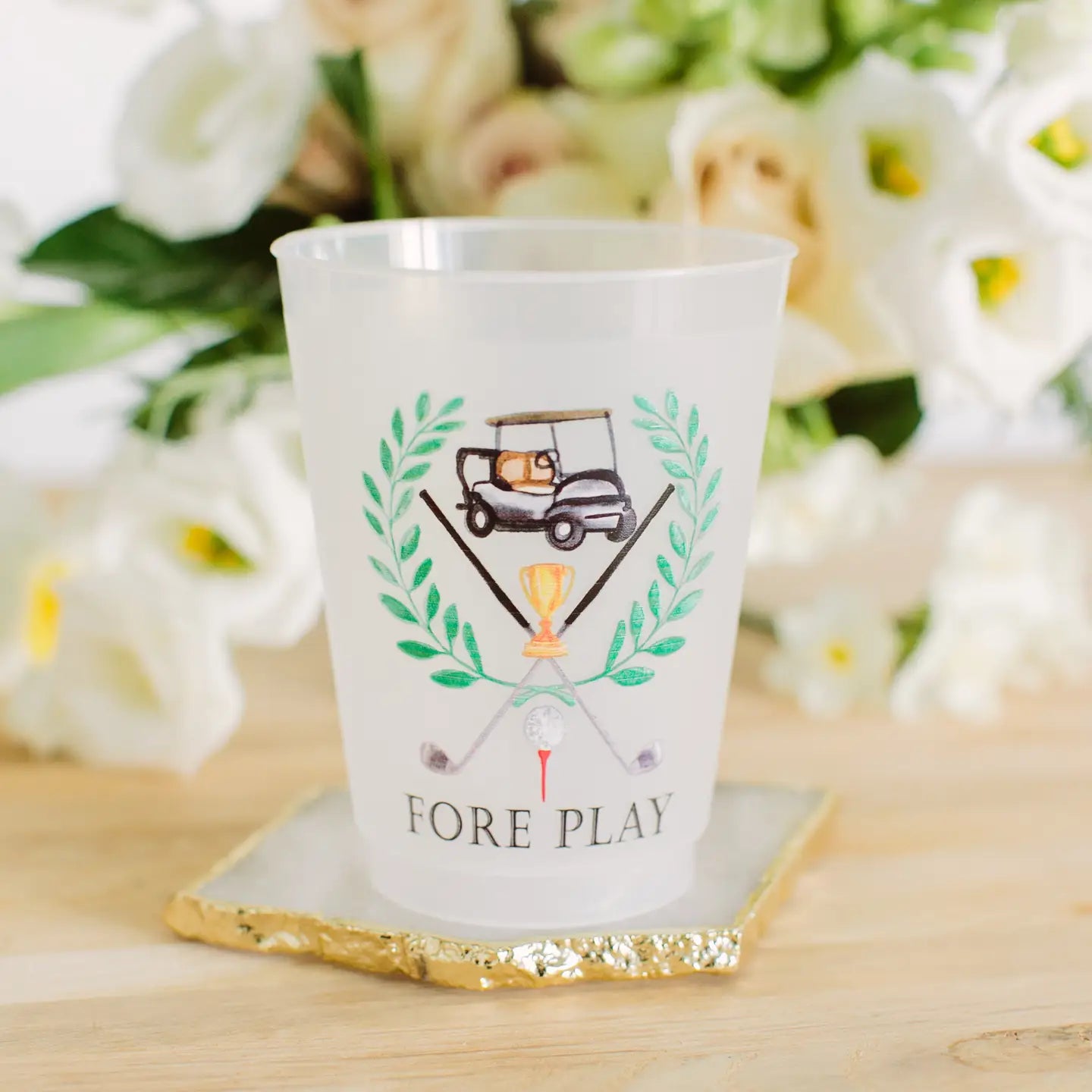 A frosted white plastic cup with a golf-themed graphic and the text 'FORE PLAY' printed on it, resting on a gold-edged square coaster on a wooden surface with a blurred floral background.